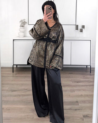 Veste kimono femme dorée et noire à motif brocart, coupe fluide avec manches larges et ceinture noire tressée, portée avec pantalon noir ample.