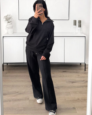 Ensemble de survêtement noir composé d’un sweat à zip à col montant et d’un pantalon larges à taille élastique avec cordon de serrage, conçu dans un tissu doux et confortable. L’ensemble présente une coupe ample et décontractée, avec des lignes verticales discrètes sur le pantalon.