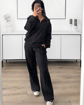 Ensemble de survêtement noir composé d’un sweat à zip à col montant et d’un pantalon larges à taille élastique avec cordon de serrage, conçu dans un tissu doux et confortable. L’ensemble présente une coupe ample et décontractée, avec des lignes verticales discrètes sur le pantalon.