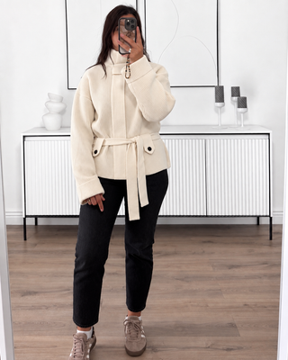 Gilet en maille écru côtelée à col montant, manches oversize et ceinture, porté avec un jean noir.