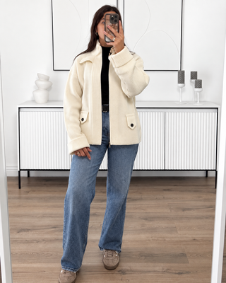Gilet en maille écru côtelée à col montant, manches oversize et ceinture, porté avec un jean bleu.