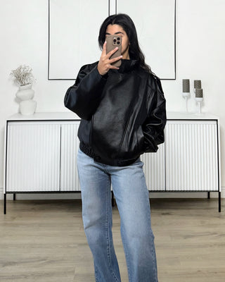 Femme portant une veste bomber oversize noire effet cuir, avec un jean bleu clair, prenant une photo dans un intérieur minimaliste.